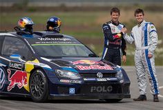 two men standing next to a car on a race track with another man in the background