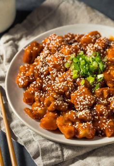 a white plate topped with meat covered in sesame seeds and green onions next to chopsticks