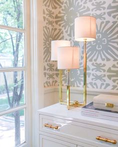 two lamps sitting on top of a white dresser in front of a window with an ornate wallpaper