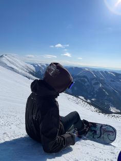 a person sitting in the snow with a snowboard on their feet, looking at mountains