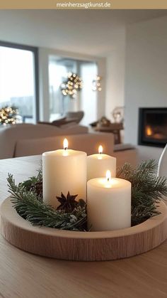 three lit candles in a wooden bowl on a table with greenery and pine cones