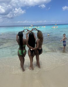 three women in bikinis standing on the beach with their hands in the shape of a heart