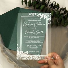 a person holding up a wedding card in front of a bouquet of flowers and greenery