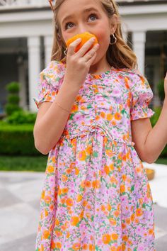 Introducing the Mini Katie Dress, the perfect garden party attire for your little flower! Made from vibrant orange, pink, purple, green, and white floral-print fabric, this dress features a darling sweetheart neckline adorned with charming ruffle detailing. With half-length puff sleeves and a knee-length skirt boasting pockets and a large ruffle hem, it's a delightful blend of style and comfort for her summer adventures. Available in sizes 12-18 months, 2T, 3T, 4-5, 6-7, 8, 9-10, 11-12, + 13-14! Playful Floral Spring Dress, Short Sleeve Ruched Floral Dress For Garden Party, Ruched Floral Dress With Short Sleeves For Garden Party, Spring Dress With Ruffle Hem And Sweetheart Neckline, Playful Ruffle Dress For Brunch, Playful Ruffled Floral Dress For Spring, Spring Dress With Ditsy Floral Print And Sweetheart Neckline, Playful Pink Dress For Brunch, Playful Short Sleeve Dresses For Brunch