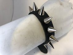 Classic gothic/punk spike bracelet with one row of 1/2” cone spikes. Made with black hand dyed vegetable tan leather.- Spikes screw in and aren’t that sharp. - Spikes made of nickel plated brass - All other hardware nickel plated- Adjustable, strap has five holes for custom sizing- Has five holes for custom sizing (Smaller bracelets have 4)I’m also able to make this in other sizes or custom colors like red, brown, pink and oxblood and in custom sizes. If you’re interested please ask! Edgy Silver Studs Jewelry, Black Spiked Edgy Bracelets, Punk Leather Bracelet With Studs For Festivals, Rock Style Spiked Jewelry For Parties, Punk Style Studded Leather Bracelet For Festivals, Edgy Black Spiked Bracelets, Rock Style Spiked Party Jewelry, Rock Style Party Jewelry With Spikes, Punk Jewelry With Spikes For Concerts