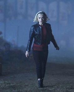 a woman is walking in the fog wearing a black leather jacket and red sweater with her hair pulled back