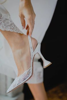 a woman wearing white high heel shoes with lace on the heels and her hand holding onto the shoe