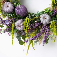 a wreath with purple flowers and greenery hanging from the front of a white wall