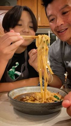 two people sitting at a table eating noodles with chopsticks