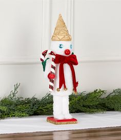 a white snowman with a gold hat and red scarf standing in front of evergreen branches