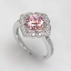 The Platinum Plated Sterling Silver Ring, Cushion Super Light Pink CZ Stone Milgrain Vintage 2 carat Engagement Ring, Pink Diamond Simulant Ring Cocktail Ring (Size 5 to 9) is nothing short of breathtaking. With its vintage-style vibe, it showcases an eye-catching light pink cushion cut cubic zirconia encircled by sparkling round cut accents set in milgrain edge finish petals being formed a heart shape. It would be a sensation at a garden party and a lovely complement to your little black dress Pink Cluster Ring With Round Cut For Gift, Elegant Pink Cluster Promise Ring, Classic Pink Cluster Ring With Halo Setting, Classic Pink Cluster Ring With Prong Setting, Classic Pink Cluster Ring With Center Stone, Classic Pink Halo Ring With Gemstone, Classic Pink Gemstone Halo Ring, Pink Cluster Ring With Round Cut For Anniversary, Pink Cluster Ring With Center Stone For Anniversary