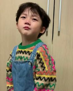 a young boy standing in front of a wall wearing overalls and an ugly sweater