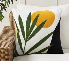 a close up of a pillow on a couch with a plant in the corner and a wicker basket next to it