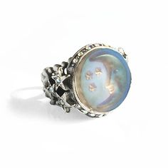 The subject of this ring is the moon and stars expressed in iridescent aurora satin glass. Light moves through the glass every way you position your hand, opalescent and glowing in soft hues of blue, purple, aqua, peach and lavender. It is surrounded by an aurora crystal border with three carved stars on both sides which join it to the ring shank, a tapering, comfortable pierced band that may be adjusted to fit sizes 6 through 9. Choice of metal finish in burnished bronze or antique silver. Made