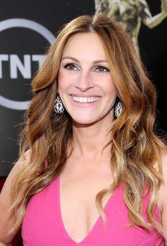 a woman in a pink dress smiling at the camera with long hair and big earrings