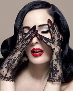 a woman with black hair and red lipstick holding her hands up to her face while wearing lace gloves