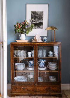 a glass cabinet with plates and cups on it in front of a blue wall that has a framed photograph above it