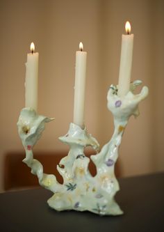 three candles are lit on a table in front of a wall behind them is a figurine with flowers and leaves