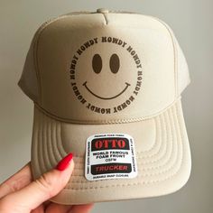 The perfect hat for beach days and country concerts alike! This hat is comprised of an Otto brand foam trucker hat in color khaki with a brown howdy smiley face design. Trendy Beige Trucker Hat, One Size Fits Most, Trendy Beige Trucker Hat One Size Fits Most, Trendy Brown Trucker Hat For The Beach, Trendy Brown Summer Trucker Hat, Trendy Summer Baseball Cap With Smiley Face, Trendy Beige Snapback Trucker Hat, Trendy Brown Snapback Hat For Summer, Trendy Smiley Face Baseball Cap For Summer, Summer Trucker Hat With Smiley Face