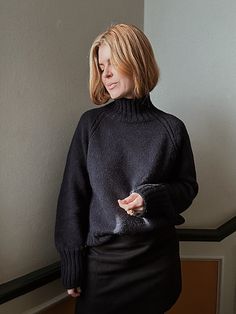 a woman standing in front of a wall wearing a black turtle neck sweater and skirt