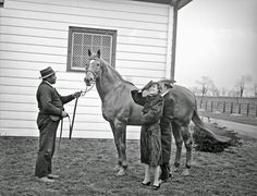 an old black and white photo of two people standing next to a horse in front of a house