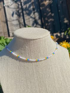 a white necklace with multi colored beads on a mannequin in front of flowers