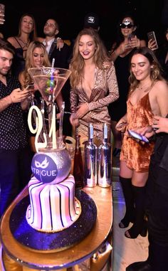 a group of people standing around a table with a cake on it and drinks in front of them