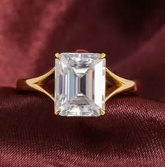 an emerald - cut diamond ring sits on top of a red velvet surface, with gold accents