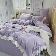 a bed with purple comforter and pillows in a white room next to a dresser