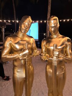 two gold statues standing in the sand with lights strung over them and palm trees behind them