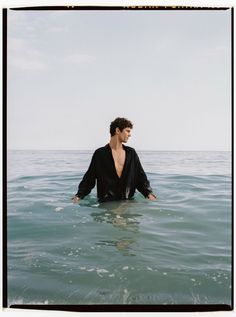 a man standing in the ocean wearing a black suit and holding his hands out to his chest