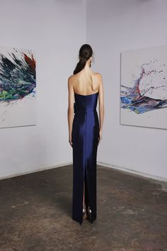 a woman in a blue dress looking at two paintings on the wall and one has her back turned to the camera