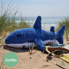 a knitted shark laying on the beach next to some books and eyeglasses