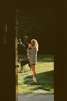 a woman holding a baby in her arms while standing next to a dog on the grass