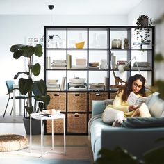 a woman sitting on a couch reading a book in a room with plants and bookshelves