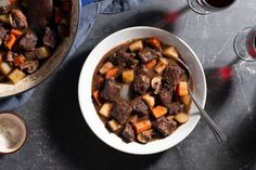 a bowl of beef stew with carrots, potatoes and gravy next to two glasses of wine