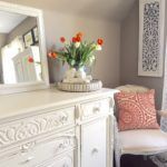 a white dresser sitting next to a window with flowers on top of it and a mirror above it