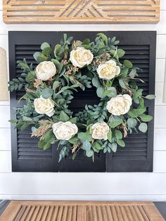 a wreath with white flowers and green leaves on a fireplace mantel in front of a black shuttered window