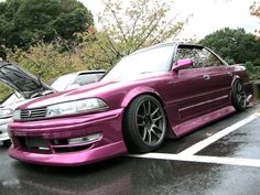 a pink car parked in a parking lot next to another car with its hood open