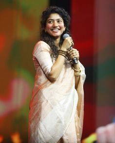 a woman holding a microphone in her right hand and wearing a white sari on stage