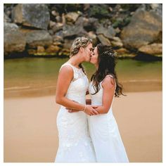 two brides kissing on the beach with rocks in the backgrouund and water behind them