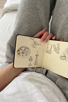 a person sitting on a bed holding an open book with drawings and food in it