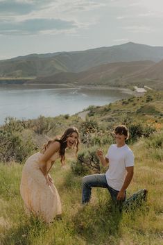 a man kneeling down next to a woman on top of a grass covered hillside near a lake