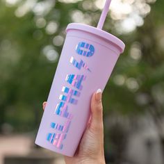 a hand holding a purple cup with pink and blue designs on the outside, in front of some trees