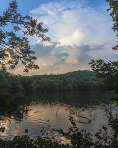 a lake surrounded by trees with clouds in the sky and sun shining down on it