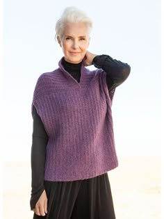 an older woman wearing a purple sweater and black pants standing in front of the ocean