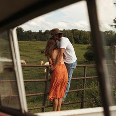 a man and woman kissing in front of a fence with cows behind them on a farm