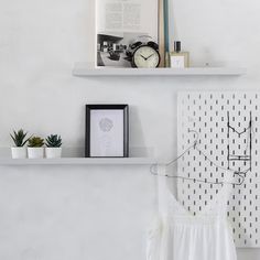 two white shelves with books and pictures on them next to a wall mounted clock, planters and other items