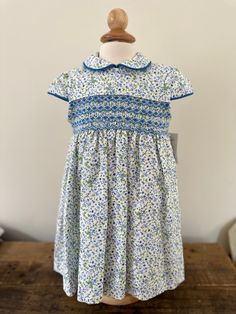 Brand: Artesania Filita Product: Nuria Composition: 100% cotton Intended Age: Toddler Description: This short sleeve, floral blue dress has a Peter Pan collar with piping (as well as on the sleeves. It is perfect for a special occasion. It fastens in the back with buttons and is fully lined. The smocked area on the chest is done in a blue pattern. Size Guide: Artisanal item- True to size. Short Sleeve Dress With Smocked Bodice For Garden Party, Garden Party Dress With Smocked Bodice And Short Sleeves, Floral Dress With Smocked Bodice For Garden Party, Fitted Floral Dress With Smocked Bodice And Short Sleeves, Spring Smocked Dress With Short Sleeves, Spring Fitted Smocked Dress With Short Sleeves, Fitted Smocked Spring Dress With Short Sleeves, Fitted Smocked Dress With Short Sleeves For Spring, Spring Cotton Smocked Dress With Lining