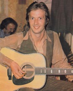 a man holding a guitar in his right hand and smiling at the camera while sitting down